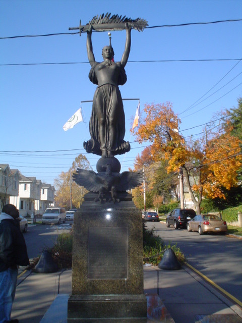 Female figure atop ball on integral plinth fronted by eagle rampant and backed by wreathed helmet, pedestal, base, four plaques