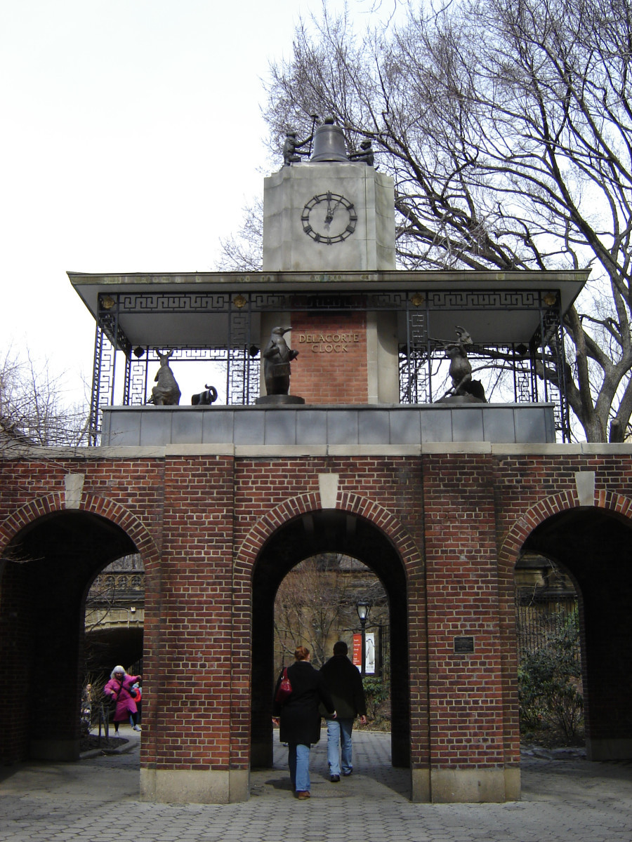 Three-tiered mechanical clock; bottom level  has six animal figures marching  around clock tower in igrillwork frame; clock faces above the animals; clock tower topped by  two monkeys and  bell; clock is on top  of arched gateway; two plaques