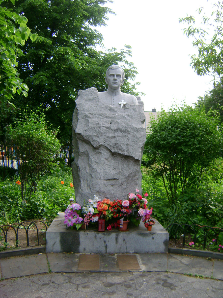 Bust emerging from a rough-hewn block, so it appears to be enshrouded up to the shoulders in a map of Poland, two plaques in pavement at front