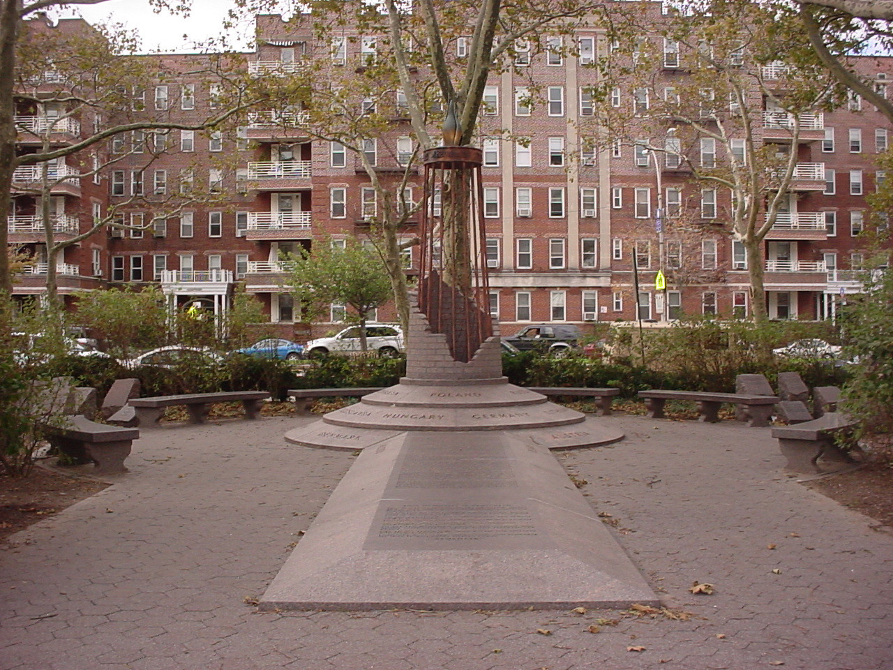 Tower consisting of brickwork partially concealing fourteen vertical bars supporting crown with beacon at top, three-tiered base, horizontal marker, approximately one hundred rough-hewn varisized vertical markers, two evergreen trees, evergreen shrubs