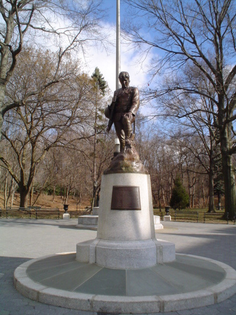 Standing figure (over life-size) contemplates a grave marked by a cross, on integral plinth, on pedestal  with plaque