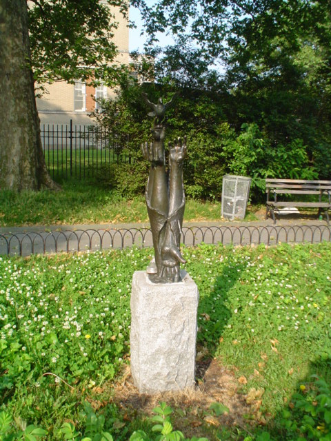 A hand pointing down supports a group of three hands pointing upward with the higher, middle hand supporting a dove with wings spread and starting to fly, on a pedestal