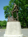 Standing figure (over life-size) contemplates a grave marked by a cross, on integral plinth, on pedestal  with plaque