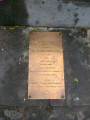 Bust emerging from a rough-hewn block, so it appears to be enshrouded up to the shoulders in a map of Poland, two plaques in pavement at front