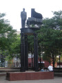 Standing figure (heroic scale) before a grand piano on a circular platform supported by three groups of three caryatids on three triform columns, integral plinth, two plaques on pedestal