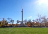 Corinthian column surmounted by standing figure (heroic scale), on pedestal with four-corner extensions bearing urns; fronted by right and left base elements each with a bas-relief; terrace with steps at front has six urns