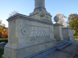 Corinthian column surmounted by standing figure (heroic scale), on pedestal with four-corner extensions bearing urns; fronted by right and left base elements each with a bas-relief; terrace with steps at front has six urns