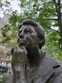 Figure (heroic scale) half-seated against a boulder, circular foot stone, earth pedestal encircled by a low wall, concentric pavement, two inscribed pavers, two plaques