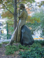 Figure (heroic scale) half-seated against a boulder, circular foot stone, earth pedestal encircled by a low wall, concentric pavement, two inscribed pavers, two plaques