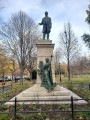 Standing figure with integral plinth on a pedestal on a three-step base; a female figure stands at the front of the pedestal in the act of writing an inscription on it