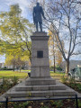 Standing figure with integral plinth on a pedestal on a three-step base; a female figure stands at the front of the pedestal in the act of writing an inscription on it