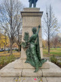 Standing figure with integral plinth on a pedestal on a three-step base; a female figure stands at the front of the pedestal in the act of writing an inscription on it