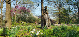 Figure (heroic scale) half-seated against a boulder, circular foot stone, earth pedestal encircled by a low wall, concentric pavement, two inscribed pavers, two plaques