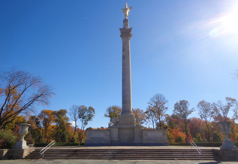 A raised paved terrace in which stands a massive limestone pedestal with sculptural reliefs. At the center of the pedestal, a Corinthian column is surmounted by a gilded bronze victory figure.