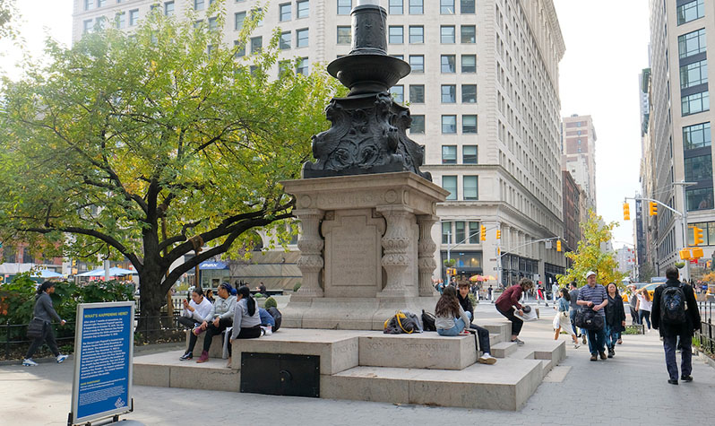 A massive stepped ornamental pink granite pedestal supporting a lavish decorative bronze cap depicting garlands and rams heads. Park-goers are sitting on the steps of the pedestal as others walk by.