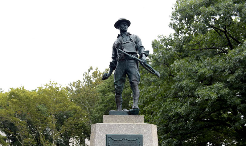 A large bronze statue on a granite base depicting a stoic, helmeted soldier in an active pose, his right hand about to hurl a grenade, and his left hand clutching a rifle with extended bayonet blade.