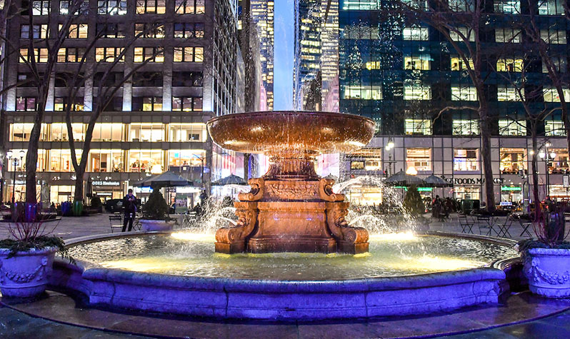 A well-lit ornamental fountain shoots water out of the sides of its basin into a reflecting pool in the early twilight.