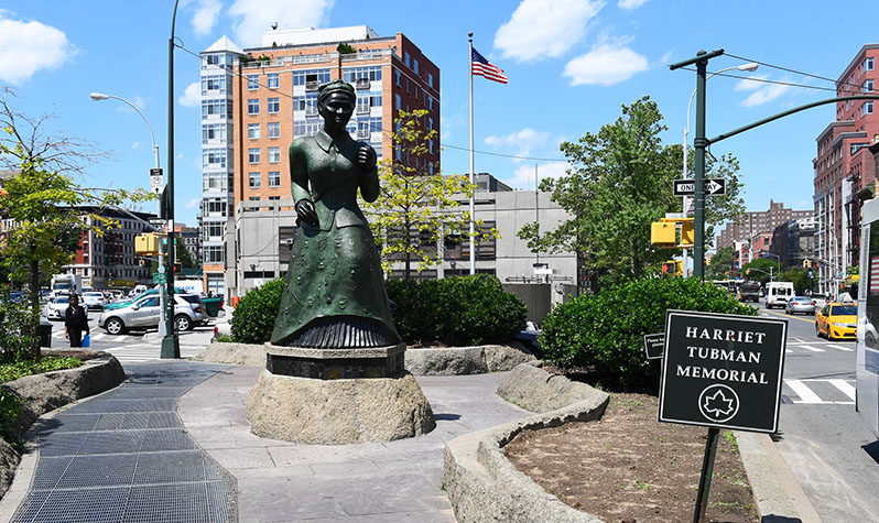 A larger-than-life statue of Harriet Tubman stands among greenery at an intersection of streets.