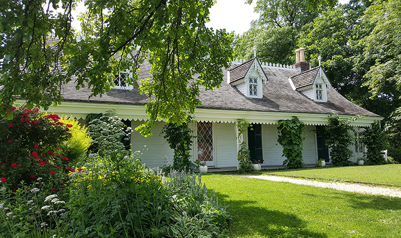 The Alice Austen House, a National LGBT Historic Site on Staten Island, is a white, one-floor cottage surrounded by a garden and a lawn with a pathway that leads to the front door.