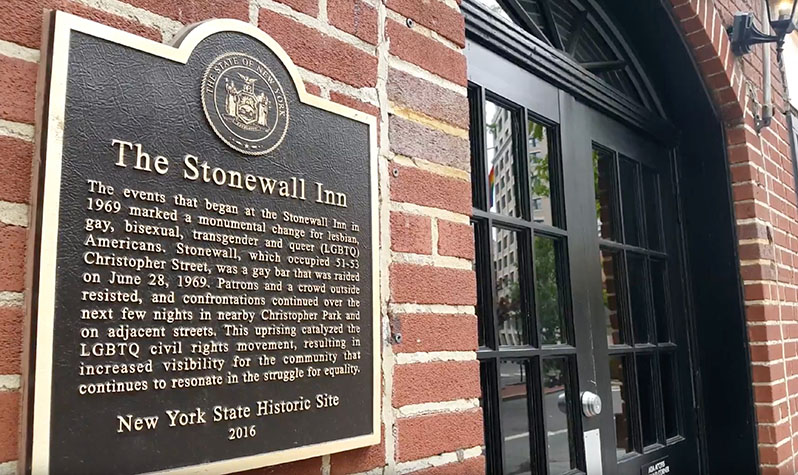 A plaque on the wall next to the door to the Stonewall Inn tells the story of the Stonewall Riots and its LGBTQ legacy. Below the history reads: New York State Historic Site, 2016.