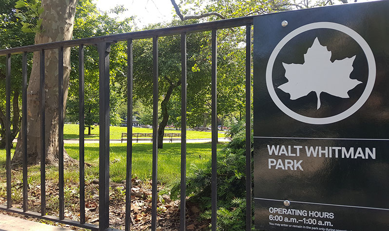 A park sign on a fence to a park area reads: Walt Whitman Park, operating hours: 6:00 a.m. to 1:00 a.m.