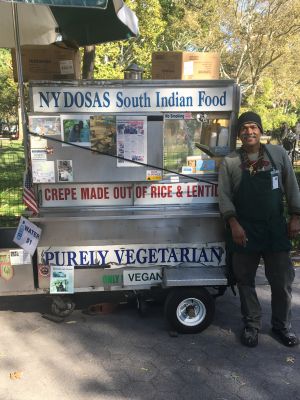 An operator stands next to a metal pushcart that reads "NY DOSAS South Indian Food" and "PURELY VEGETARIAN."