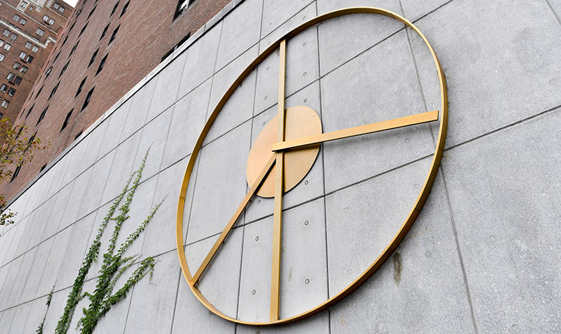 The brass kinetic sculpture on a wall functions as a clock whole hands, twice a day, shows the peace sign.