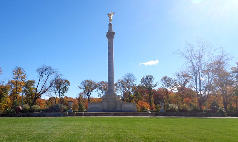 A 70-foot-tall column with a woman on top stands among a grove of trees preceded by an open lawn area.