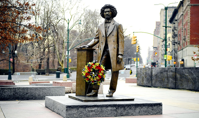 A bronze statue of Frederick Douglass standing on a platform and resting his hand on a podium. The statue is surrounded by other elements of the memorial which is contained in a cirlce.