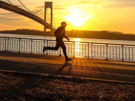 a jogger runs along pedestrian pathway that looks out to the sunset setting across from the waterway which features a view of the bridge