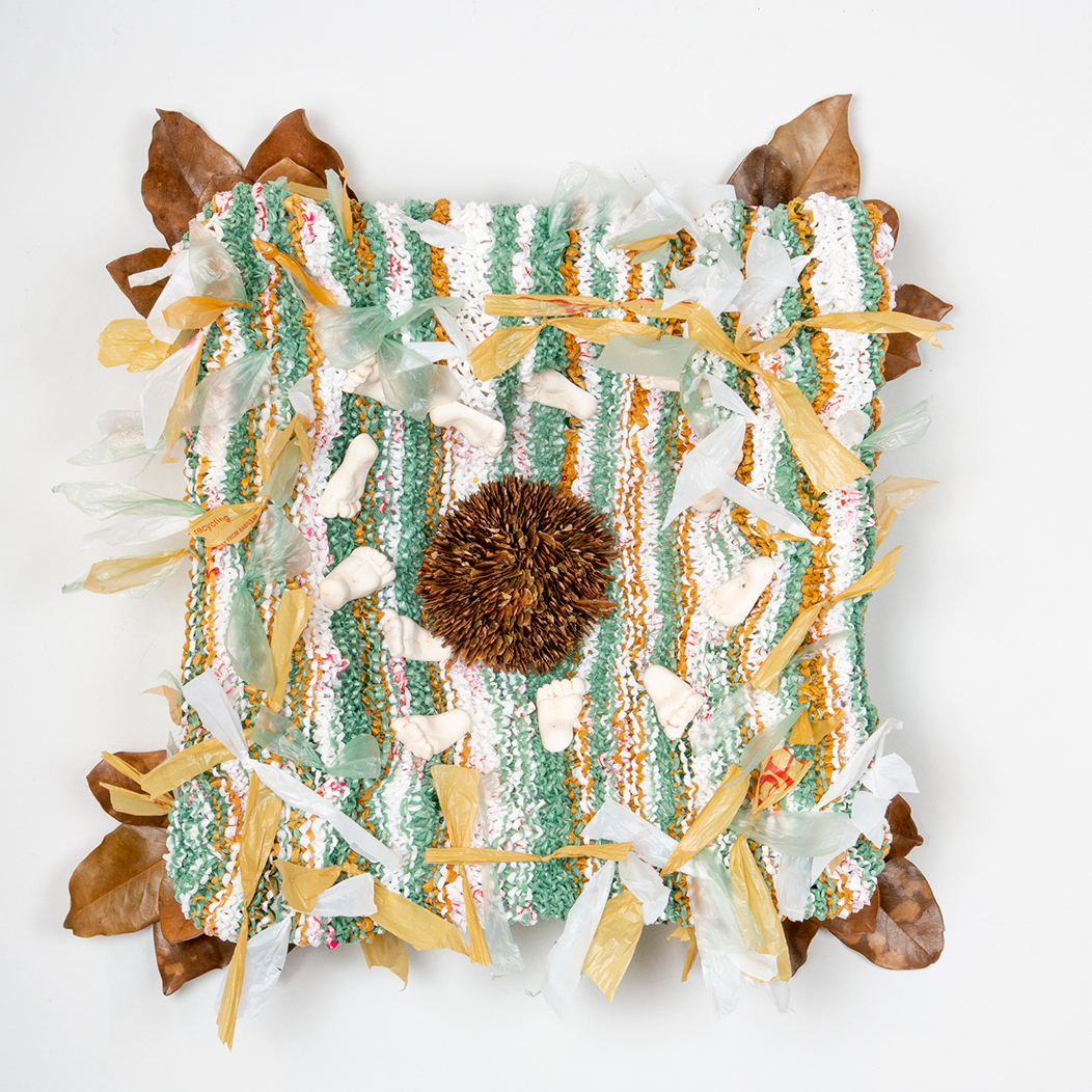 A tapestry of green, orange, red, brown, and white plastic bags woven together into a square and adorned at each corner by brown leaves with a pine cone placed in the middle