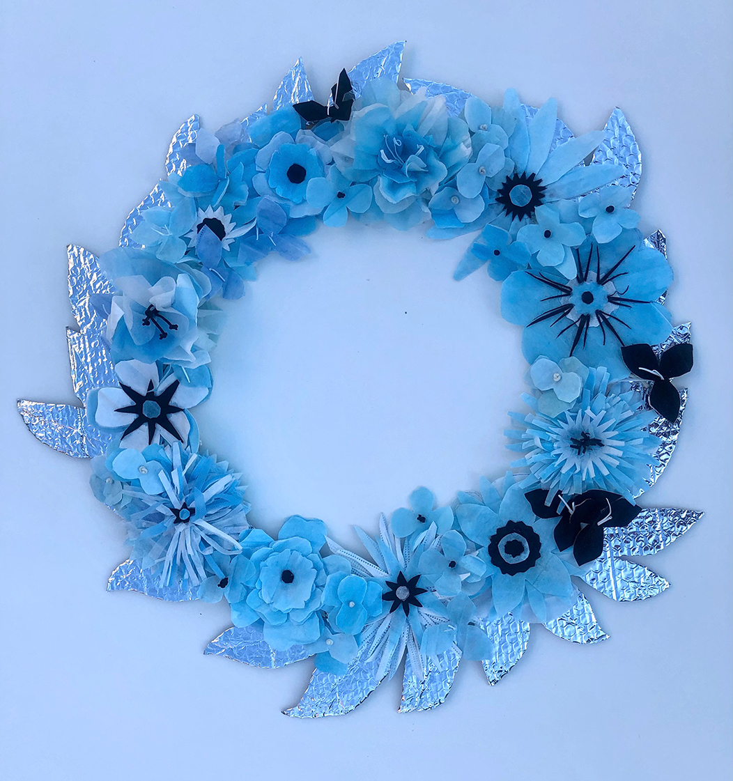 Flowers made from blue surgical masks are arranged on leaves formed from aluminum foil to form a wreath set in a blue glow