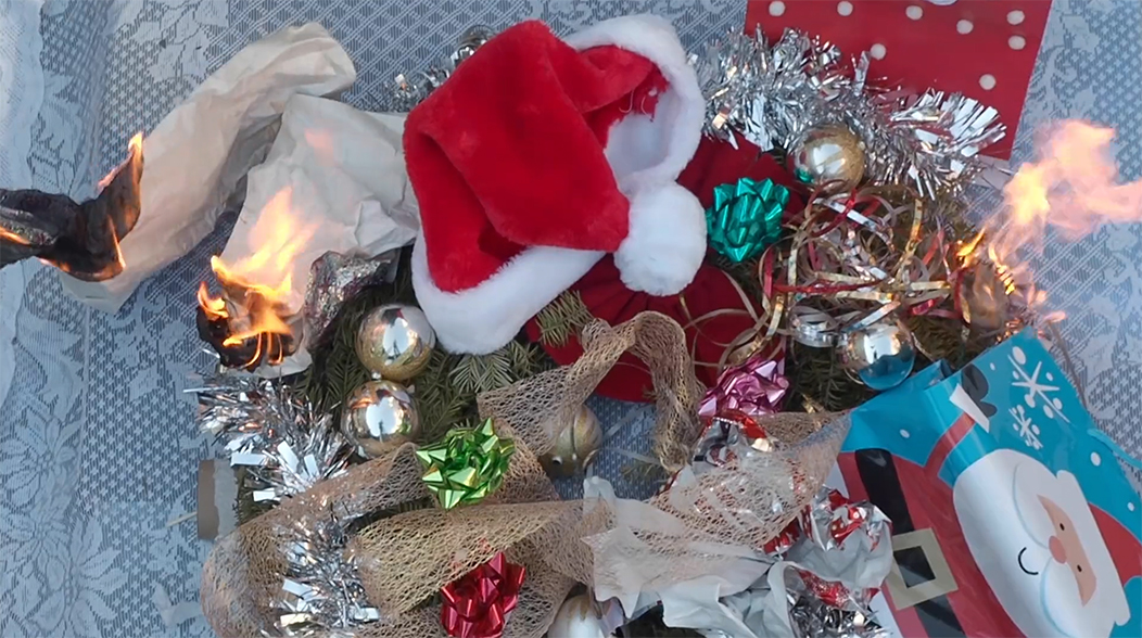 screenshot of a video of an artist whistling and humming holiday carols while placing photos on a wreath followed by a sped up video of the artist designing the wreath with holiday decor before setting it all on fire. As the wreath burns to ashes to reveal a cross, the artist states the number of acres of trees damaged by fires across the world. Young leaves are then placed at the ashes as the artist whistles a holiday carol