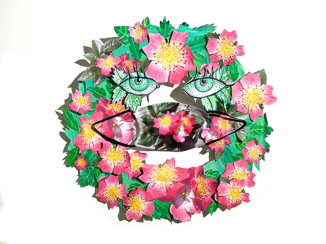 A mask decorated with painted images of pink and yellow heritage rambler roses is placed at the mouth of the wreath and just below the eyes of the wreath which is made of painted pink and yellow heritage rambler roses