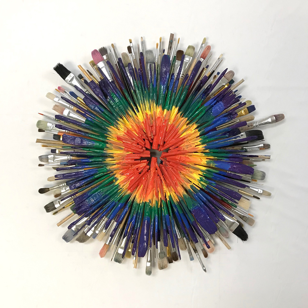 Paint brushes in various sizes, shapes, and colors are painted at the handle then arranged in a circle at the tips to form circles of colors with the innermost color being orange, yellow, green then blue