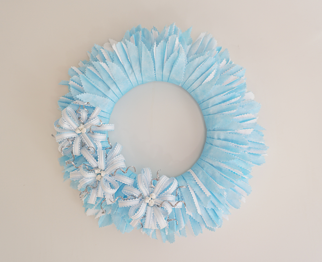 Disposable blue and white face masks are stripped and arranged to form a blue and white wreath with white bows