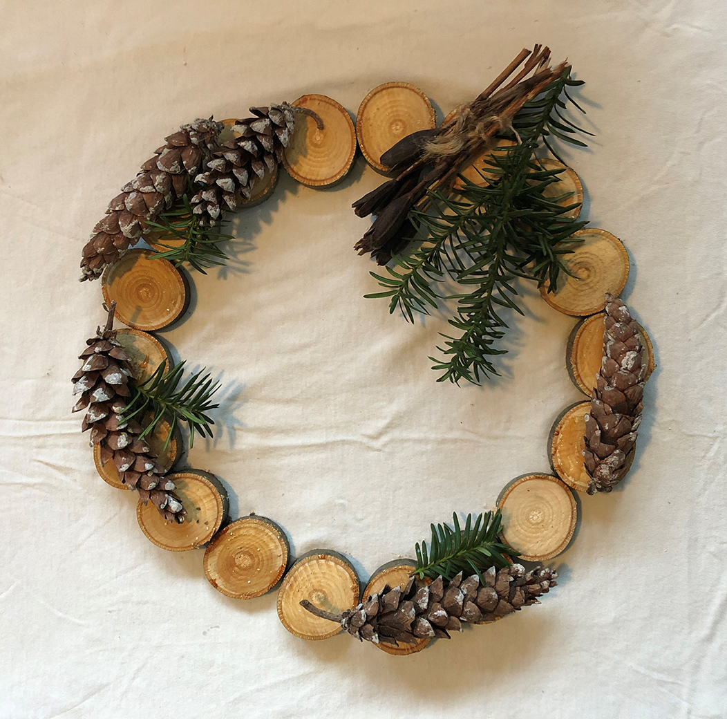 A tree branch cut into circles is arranged to form the base of the wreath which is decorated with brown yew branches, pine cones, and green iris stalks