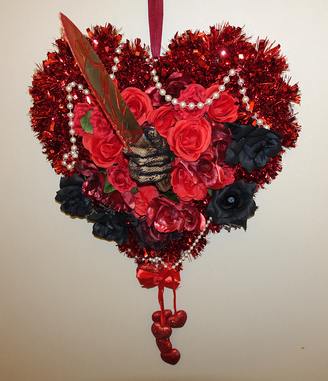 A heart shaped wreath made from red tinsel, red and black roses, pearls, and tiny hearts that hang from the bottom features a hand wearing a lace glove protruding from the center of the heart with a knife covered in blood