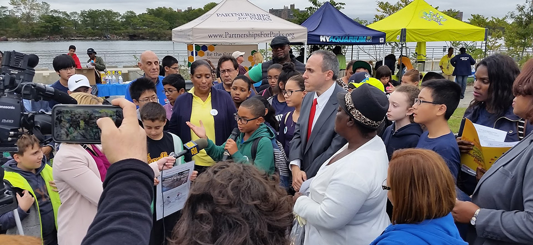 A council member, kids, and community members gather around a kid who addresses the press about the creek