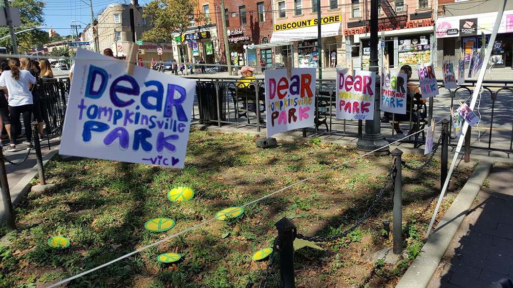 Signs that read Dear Tompkinsville Park are arranged on a clothes line along the park perimeter. Peace signs are scattered on the lawn