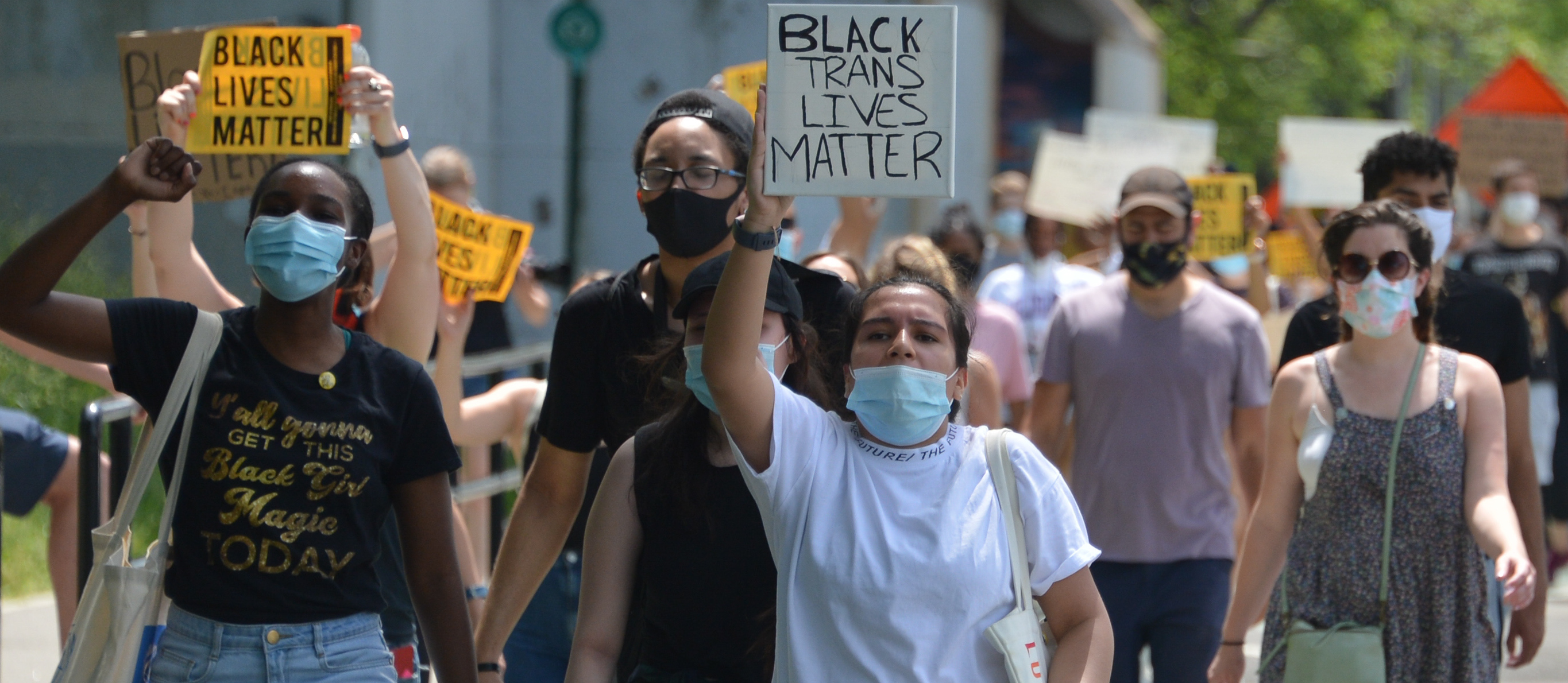 protesters holding signs that read Black Lives Matter and Black Trans Lives Matter march along a strip between a park and street