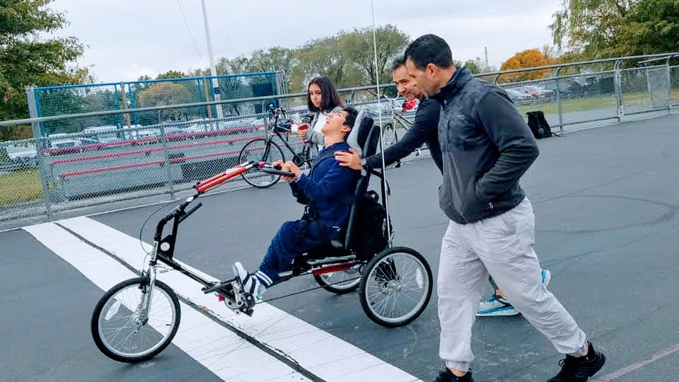 A kid is assisted with using an adaptive bike on the course in the park