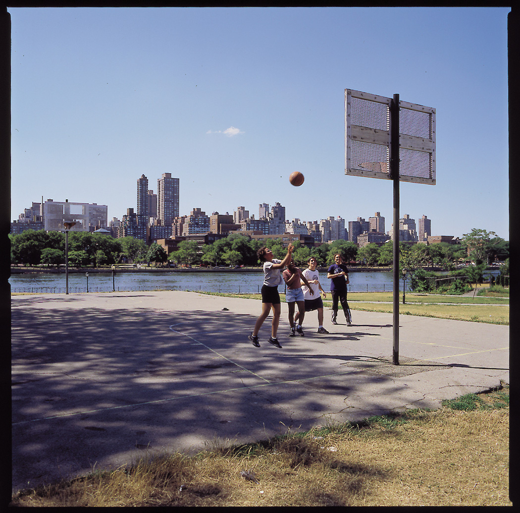 Kids play basketball at a park that features a beautiful view of the east river and the skyscrapers in Manhattan