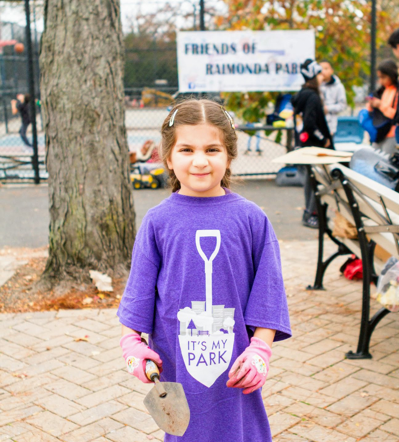 A girl wearing an It's My Park tshirt poses for a photo in front of the Friends of Raimonda Park sign