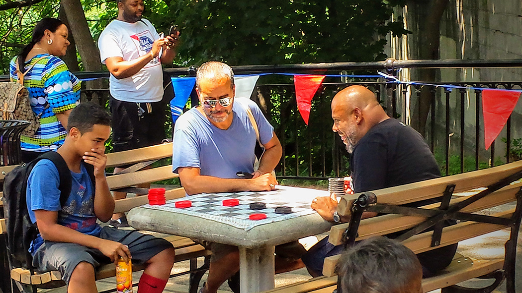 Community members challenge each other to a game of checkers and the chess and checkers table in the park