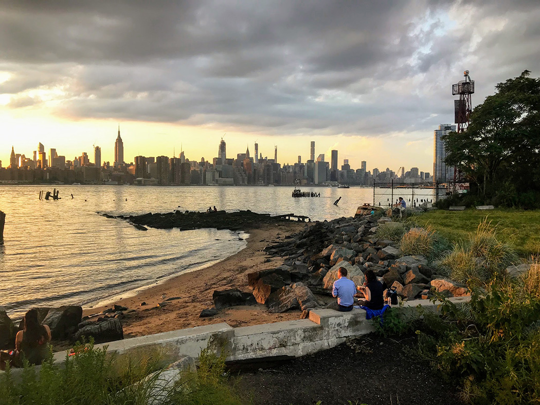 Folks enjoy the sunset while sitting at the shorefront of the park which features a view of the east river and the Empire State Building across the way