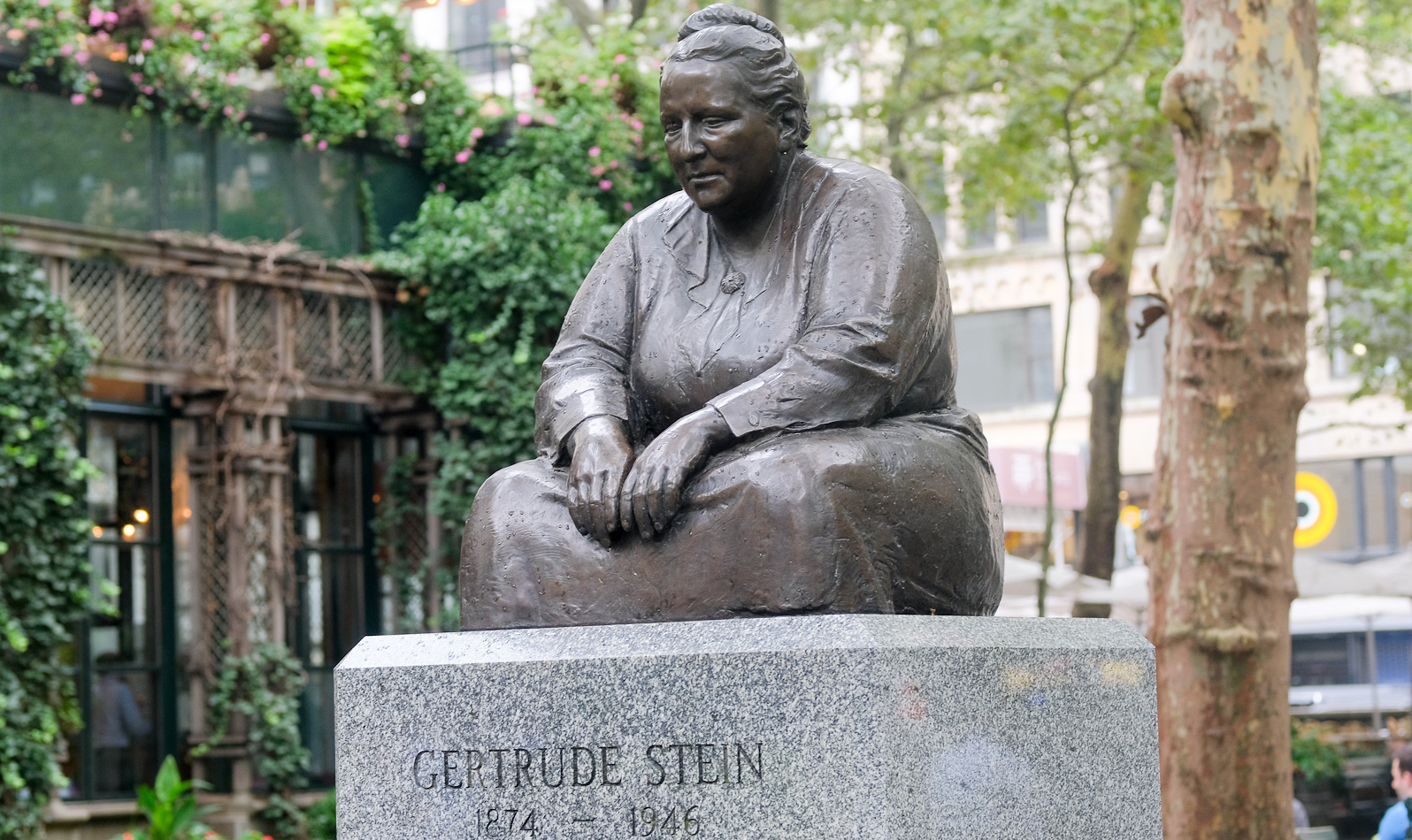 A bronze bust of a seated Gertrude Stein rests on a granite plinth in a tree-lined picnic area of Bryant Park.