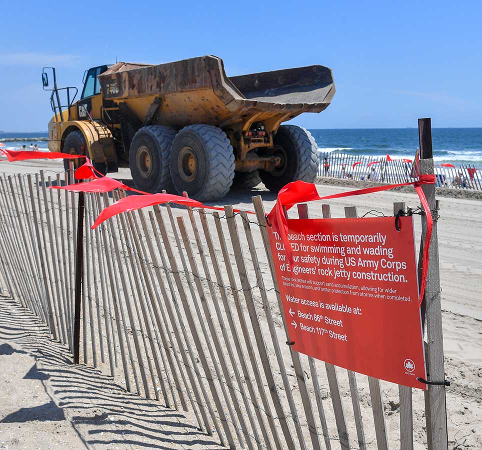 A fence with a bright red closure sign and construction tape is seen on the beach, with an earth mover truck in the background.