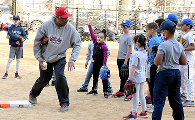 Children gather around a baseball coach teaching them how to throw a ball