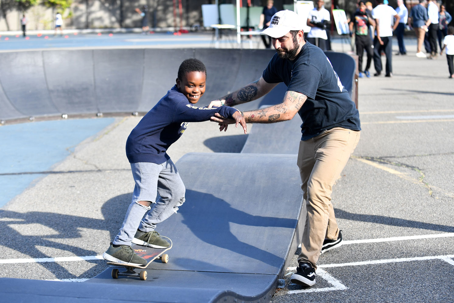 A man with multiple tattoos who looks like both a skater and coach teaching a preteen boy how to balance on a skateboard. The boy is smiling.